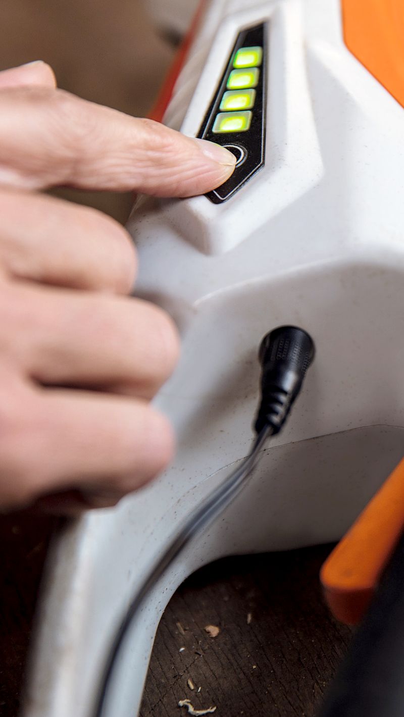 Hand dr&uuml;ckt Taste an orange-wei&szlig;em STIHL-Ger&auml;t mit leuchtender Ladeanzeige und eingestecktem Kabel auf Holztisch