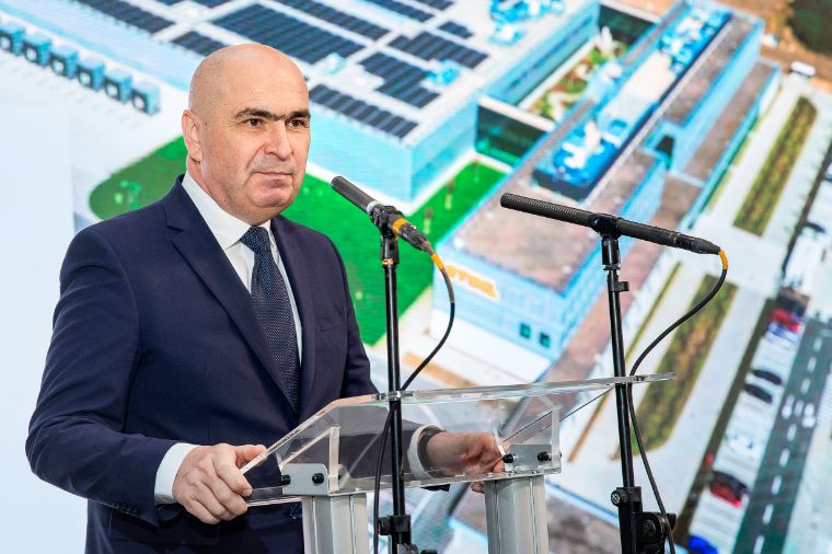 Ilie Bolojan in a dark suit speaking at clear podium with two microphones, background showing aerial rendering of modern facility