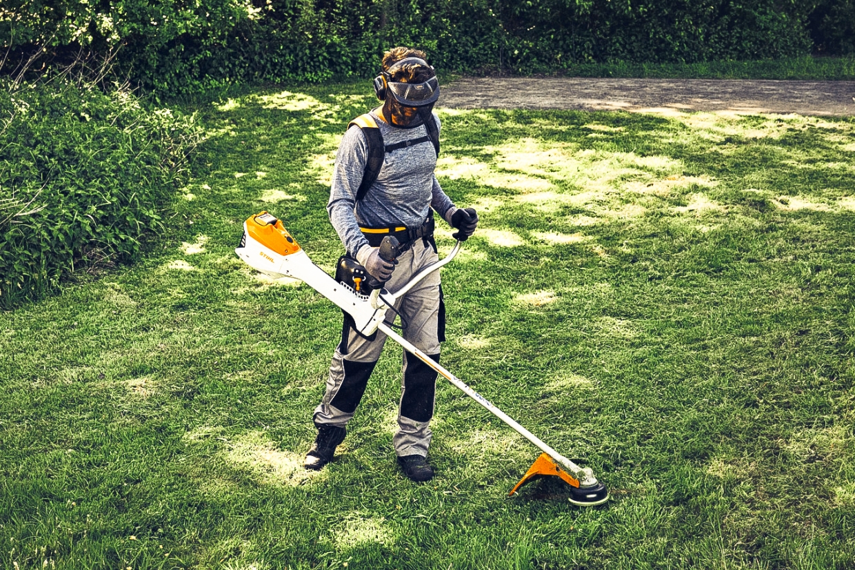 Person wearing helmet, visor and ear protection using a STIHL brushcutter on sunny lawn area beside dense garden hedge.