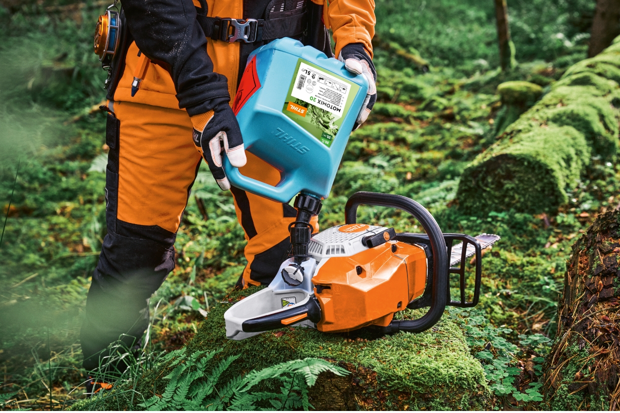 Person in orange protective clothing refuelling a STIHL chainsaw with MotoMix canister on moss covered log in forest