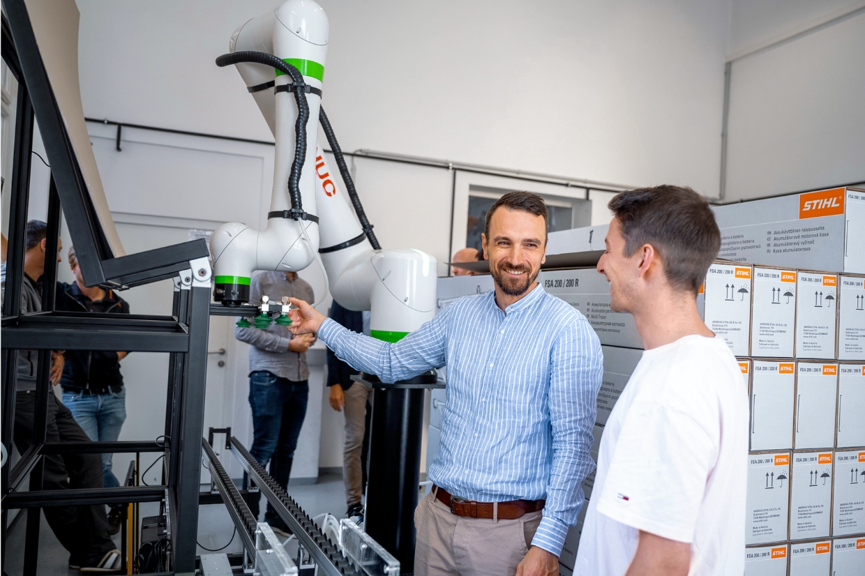 Two employees talking beside collaborative robot arm at conveyor line, stacks of STIHL product cartons arranged in background