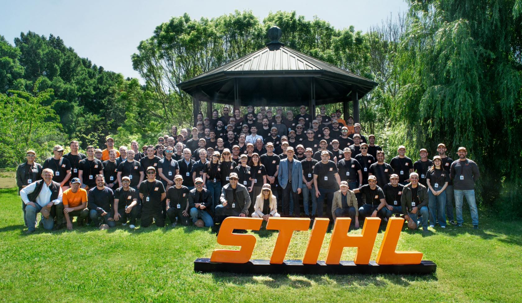 Large STIHL employee group in park before pavilion, all wearing company clothing and badges, orange STIHL sign in front