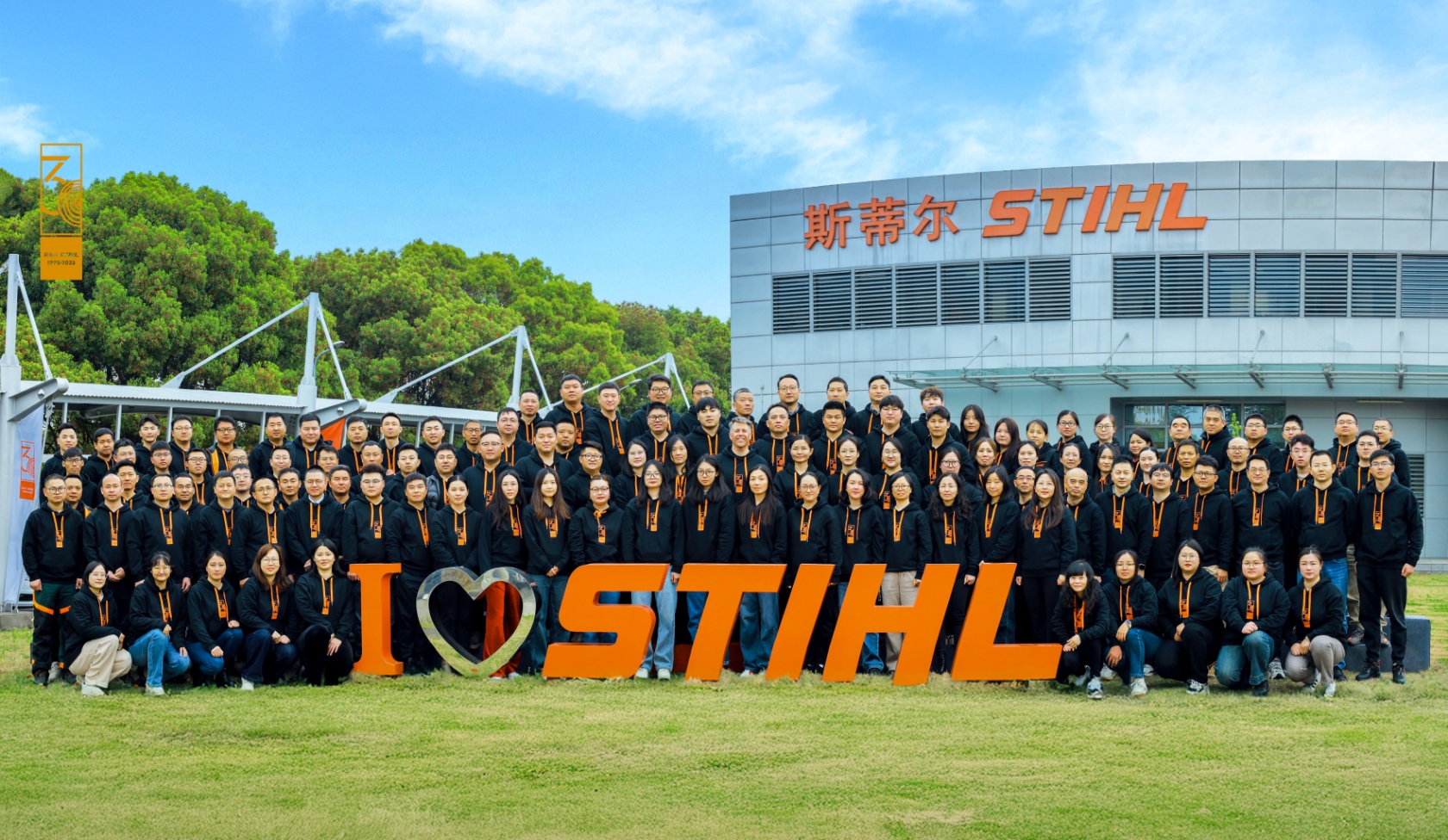 Large STIHL workforce in front of company building with logo, all in black hoodies with orange cords standing on green lawn