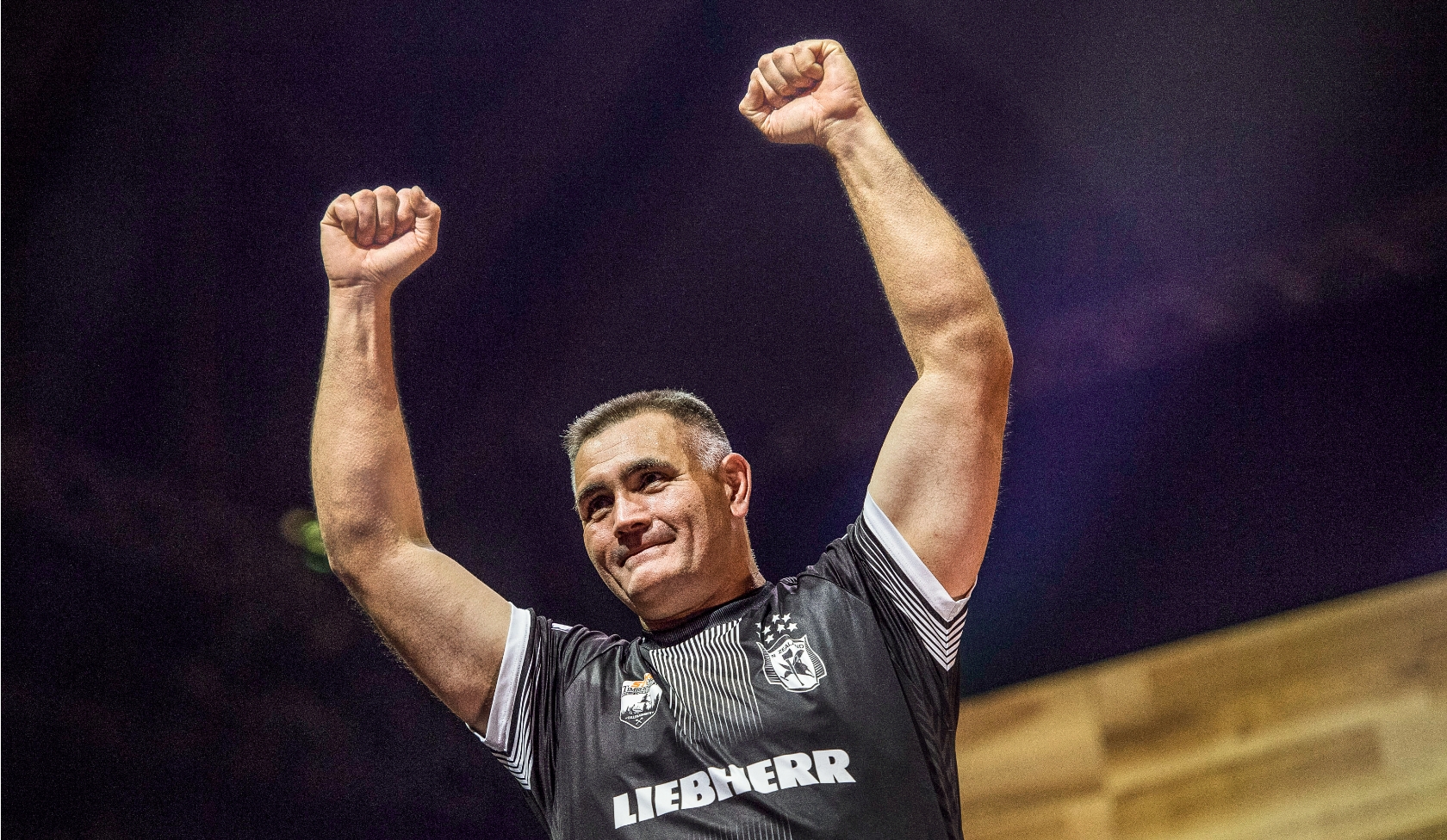 Timbersports athlete in dark jersey raising both arms on lit stage, cheering spectators fading into dark arena background