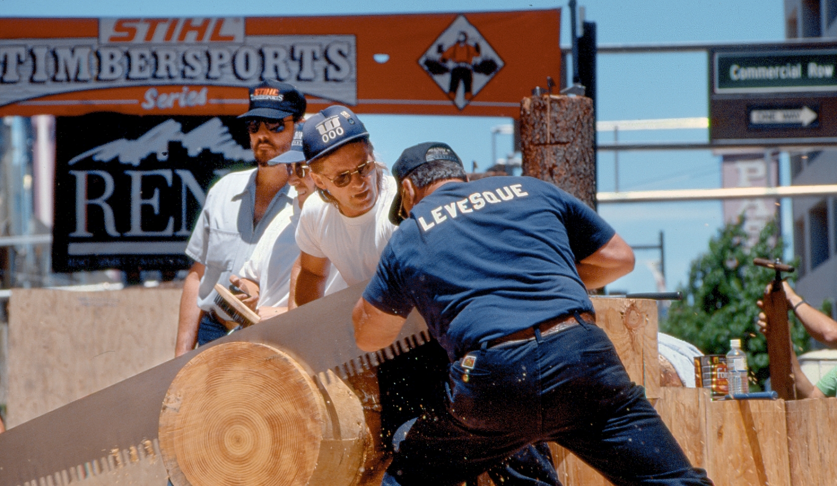 Early STIHL Timbersports scene with athletes using a two man saw on large log, spectators standing close beside arena table