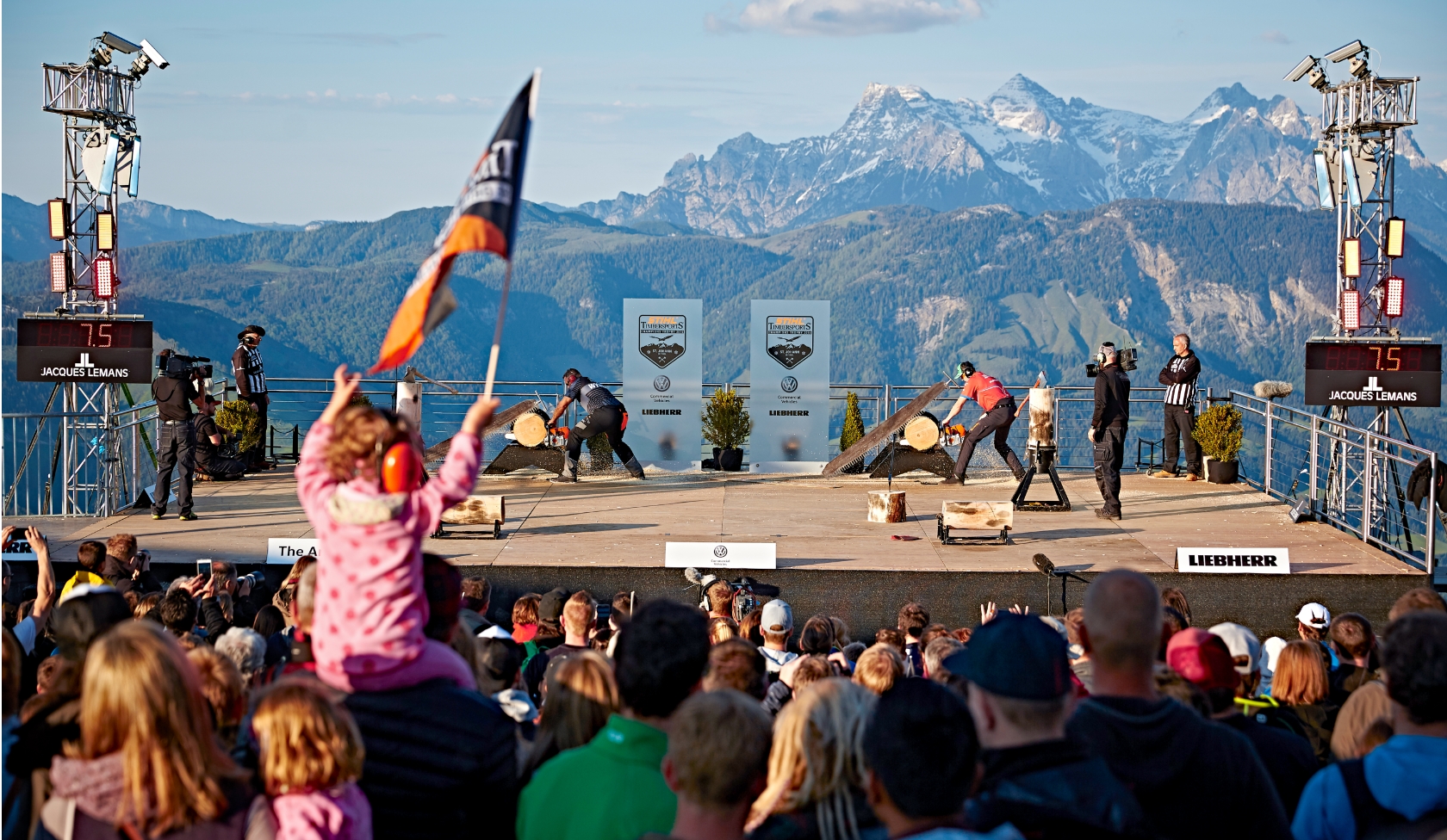 STIHL Timbersports contest on mountain stage with two sawing teams, child wearing ear protection and flag, Alps in background