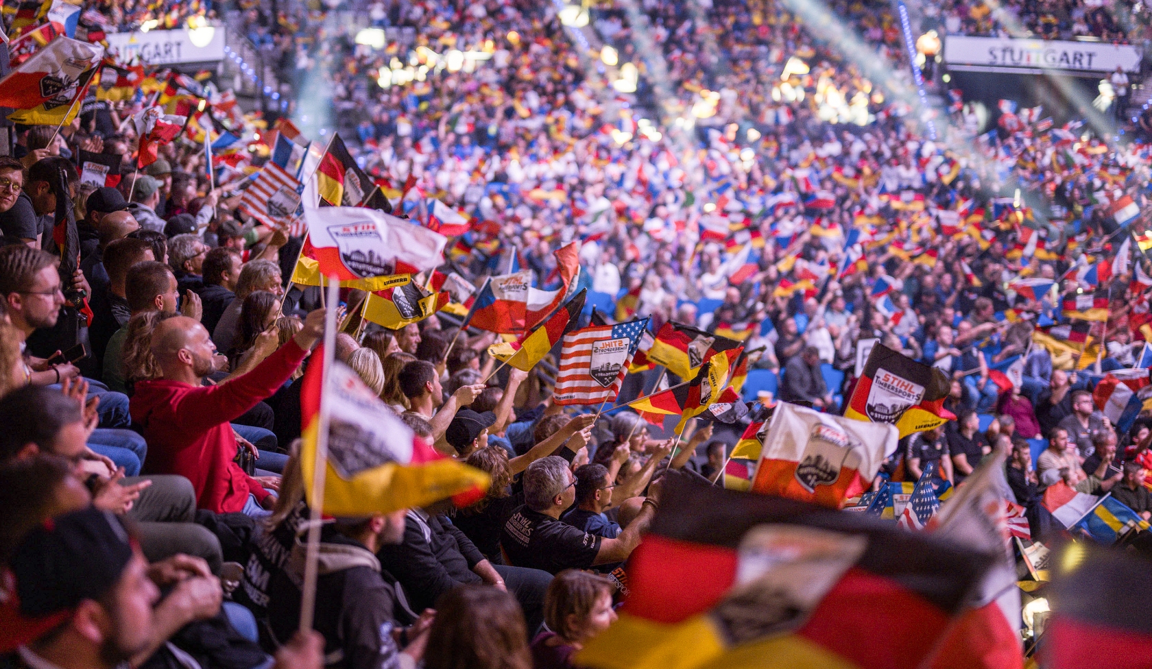 Large arena packed with STIHL Timbersports fans, hundreds waving national flags as bright spotlights sweep across stands
