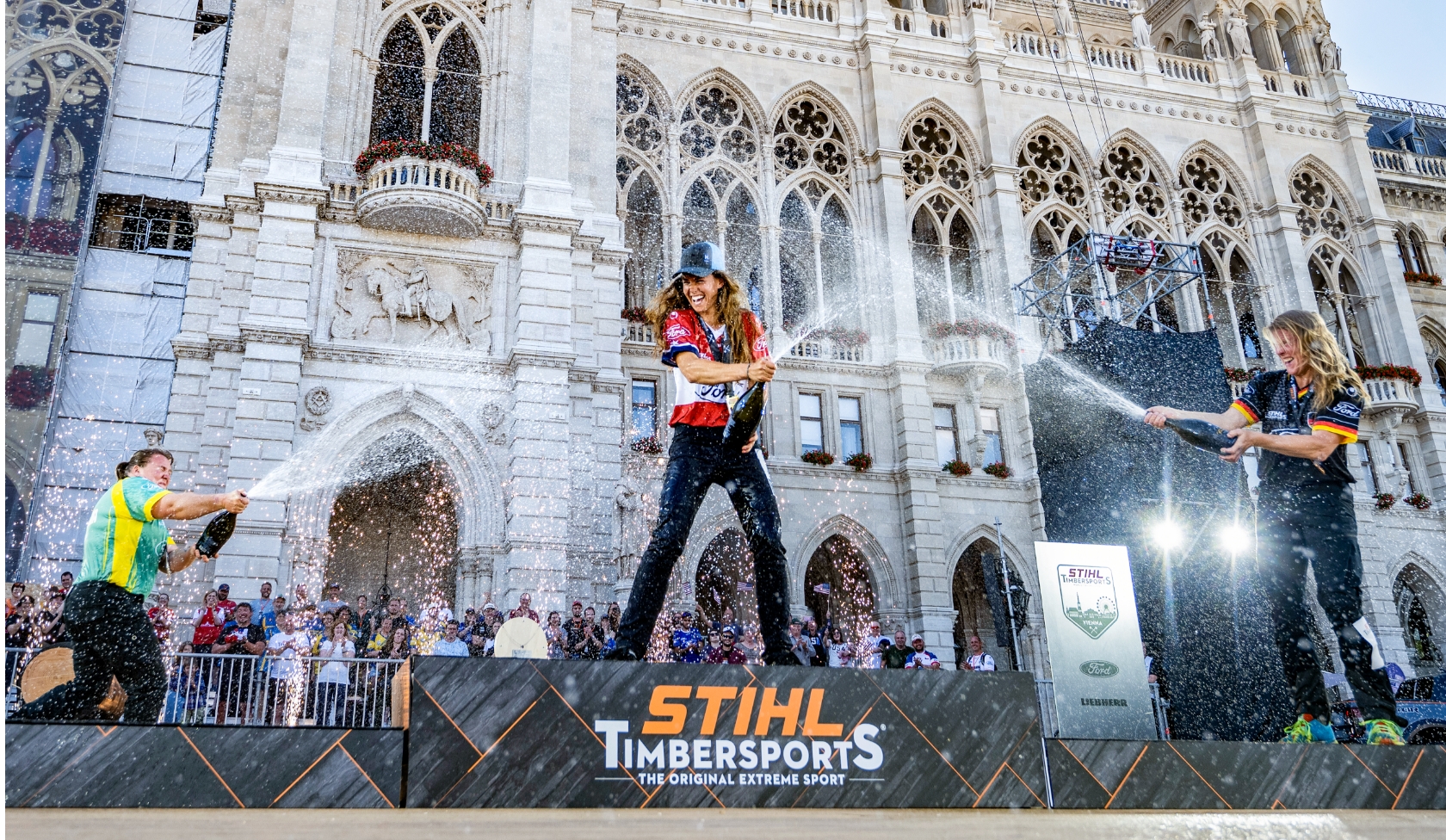 Three STIHL Timbersports athletes spraying champagne before ornate city hall facade, winners podium with logo in foreground