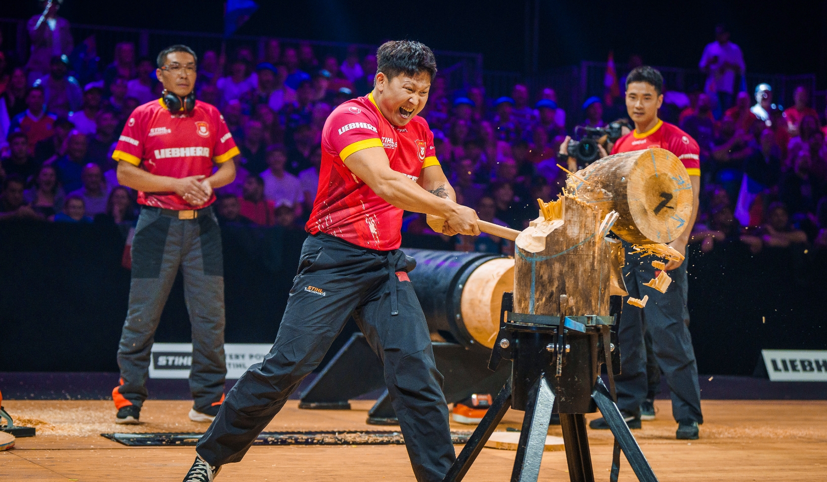 STIHL Timbersports competitor in red jersey chopping wood block with axe, teammates and audience crowding bright arena