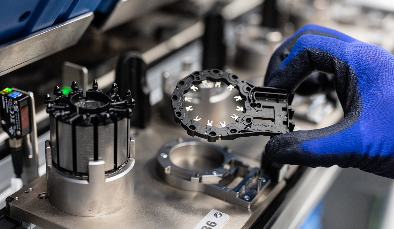 Blue gloved hand holding plastic electric motor component above assembly station with additional metal parts and sensors